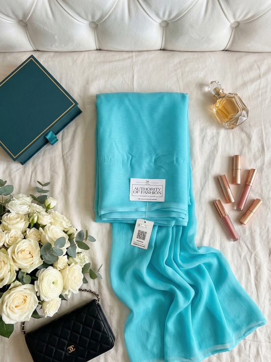 Turquoise scarf with a brand label on a white surface with flowers, book, and lip products.