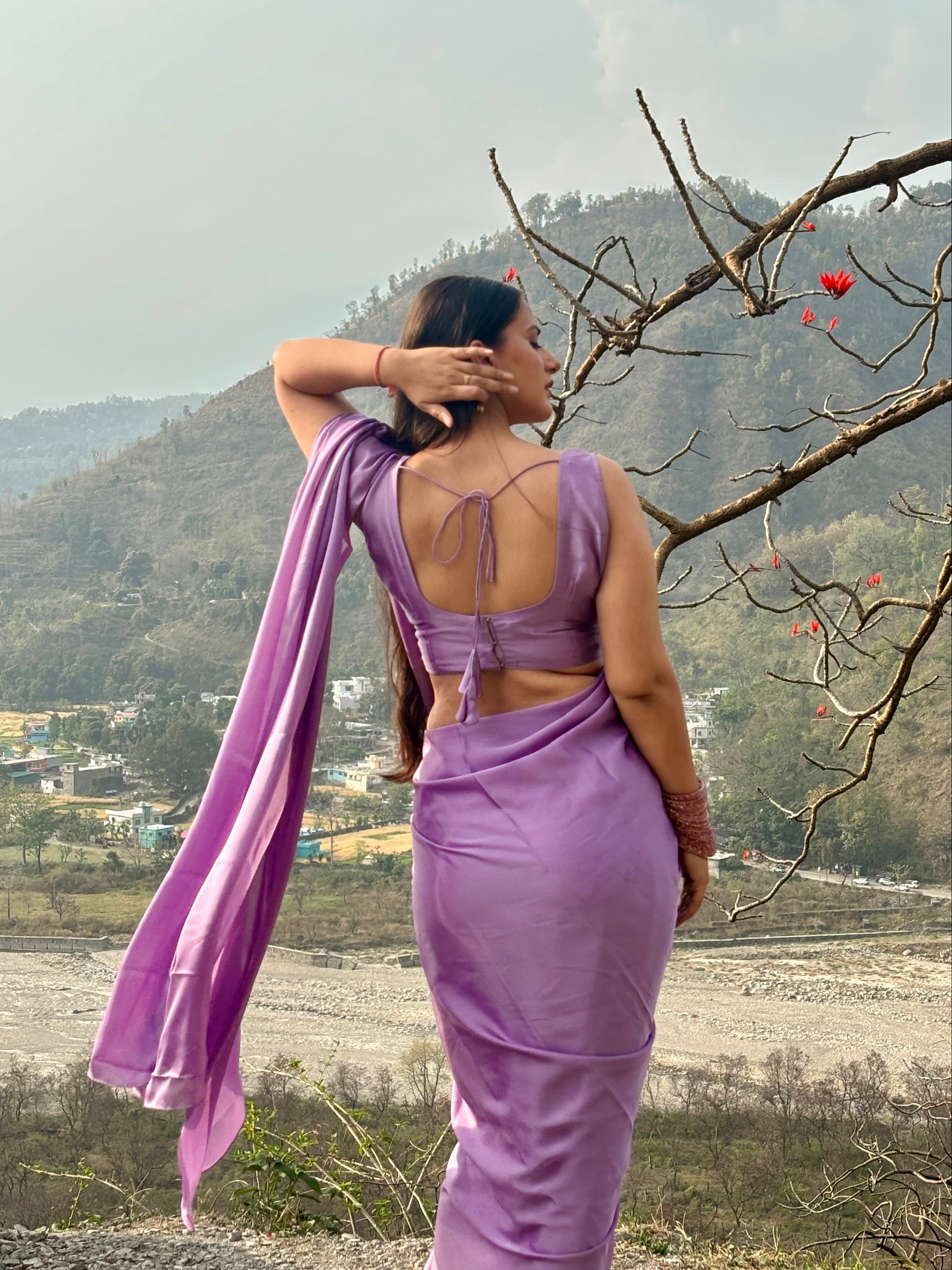 Woman in a purple saree standing near a tree with mountains in the background
