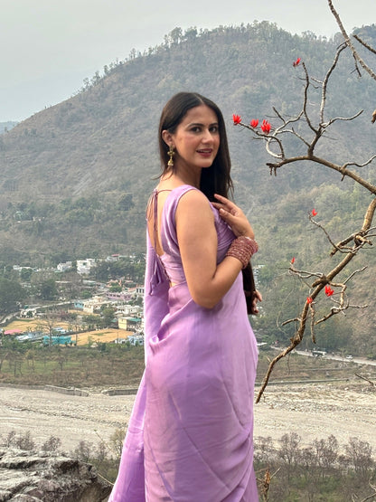 Woman in a purple saree standing outdoors with mountains and a river in the background