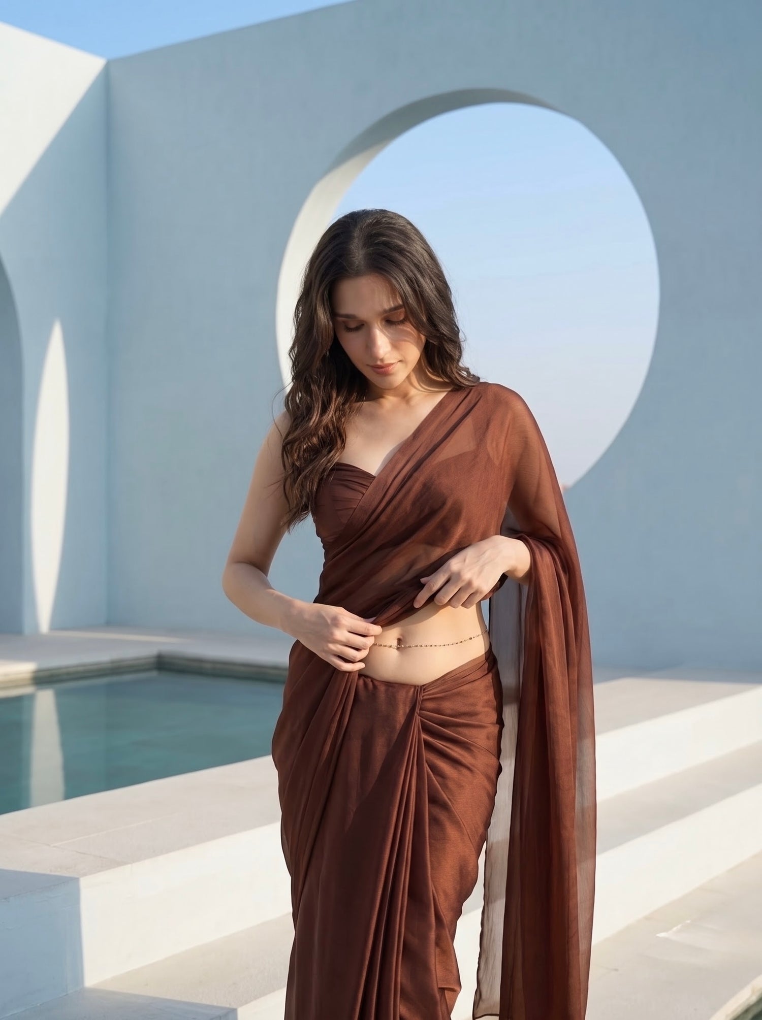 Woman in Cocoa Mirage brown chiffon saree with both hands in her hair, poolside circular archway backdrop