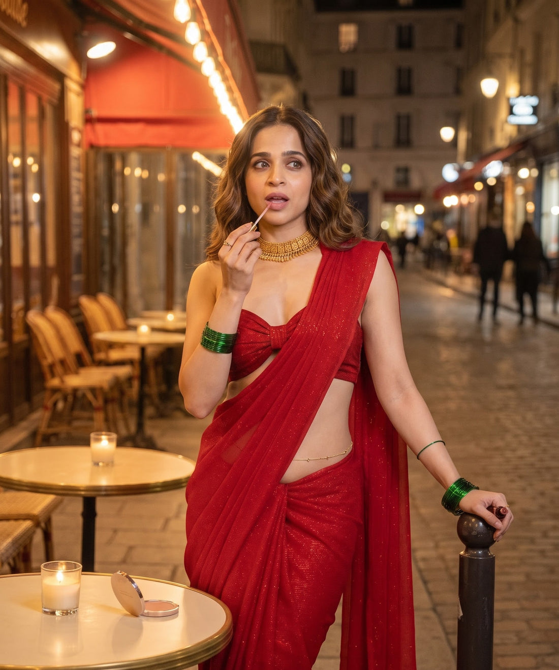 A woman wearing a shimmering red sari with a matching red sleeveless blouse stands on a city street at night, applying lipstick. The background shows a blurred Parisian-style street with cafe tables and warm lights.