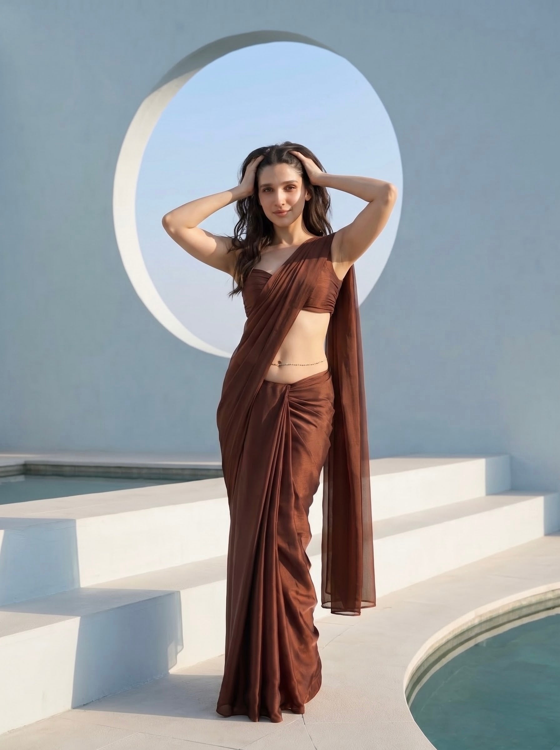 Woman in Cocoa Mirage brown chiffon saree adjusting the pallu at the waist, gold belly chain visible, poolside archway