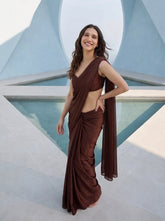 Woman smiling in Cocoa Mirage brown chiffon saree, hand on hip, framed by a triangular archway and reflecting pool