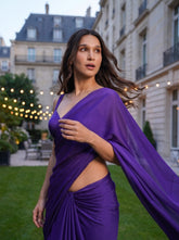 A woman wearing a flowing purple draped dress with side cutouts, standing outdoors on a grassy lawn with Parisian-style buildings and string lights in the background.