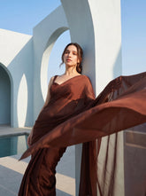 Woman in Cocoa Mirage brown chiffon saree standing between white architectural columns beside a pool