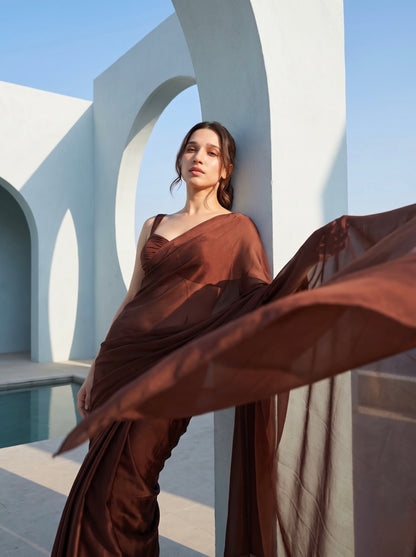 Woman in Cocoa Mirage brown chiffon saree standing between white architectural columns beside a pool
