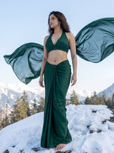 A woman stands barefoot on a snowy mountain ridge wearing a flowing dark green two-piece outfit with a halter crop top and draped maxi skirt, while sheer matching fabric billows in the wind against a mountain and sky background.
