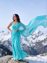 A woman wearing a flowing turquoise blue saree stands barefoot on a snowy mountain rock with snow-covered peaks in the background.