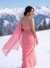 A woman wearing a pink sari and matching crop top walks through the snow with a mountain range in the background.